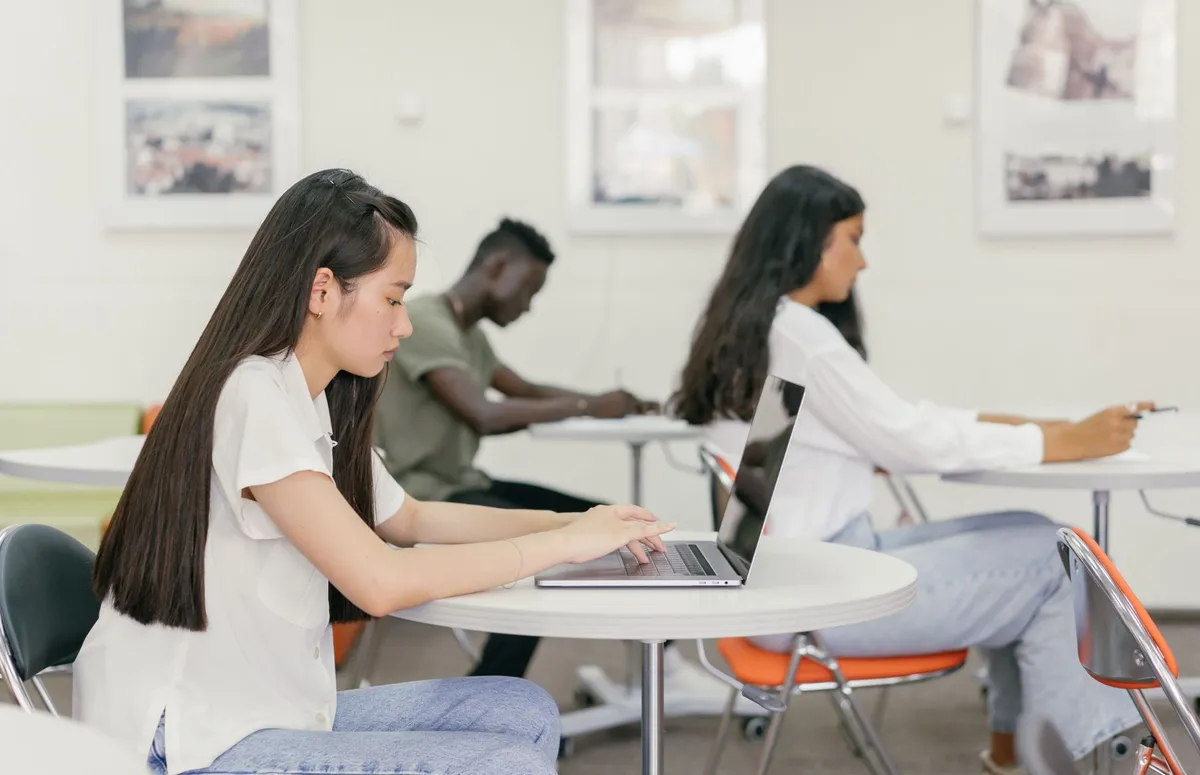 Student typing on a laptop in a bright classroom with classmates working nearby