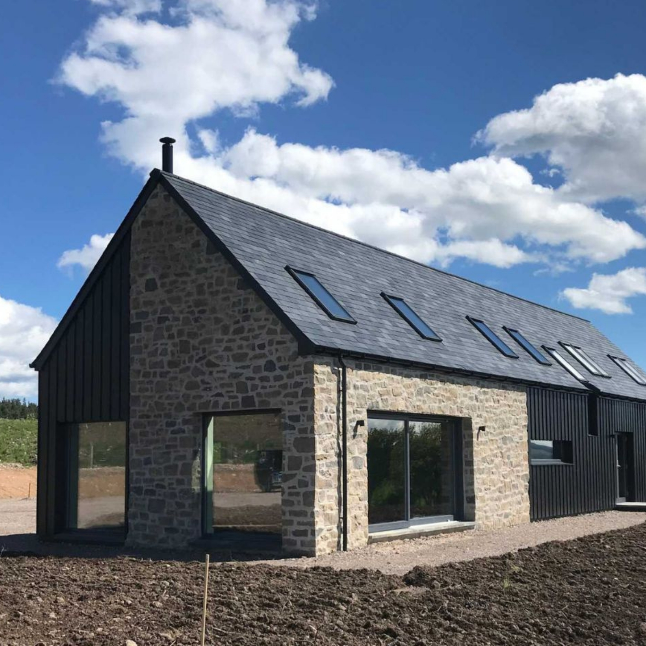 Stone built cottage with black cladding, black roof, big windows and vibrant blue sky.