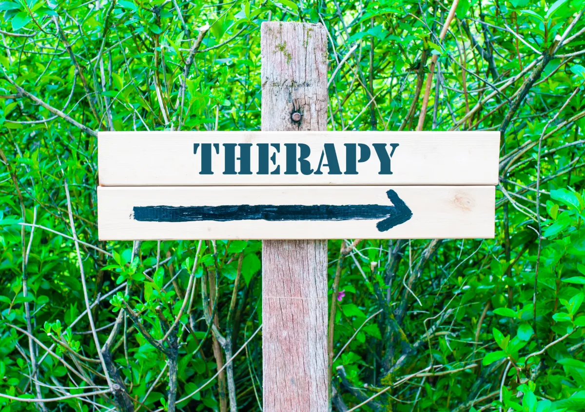 In foreground is a wooden trail sign with the word "Therapy" stenciled on top and a hand painted arrow directing the viewer right on the lower part of the sign; the background is of thick green brush.