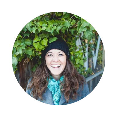Head shot of Jessica Jo (CEO of company); vibrant green foliage in background with Jessica Jo pictured in a black beanie with a teal scarf and gray peacoat. Bright laughing smile and brown curly hair. 