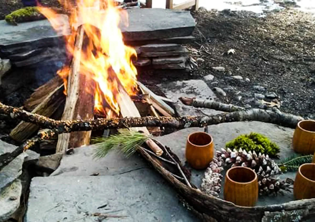 Picture of newly made fire (indicated by the logs still being visible in a tent fashion; stone seating surrounding with wooden cups, sticks, pine boughs, pine cones, and moss placed in a circular intentional manner.