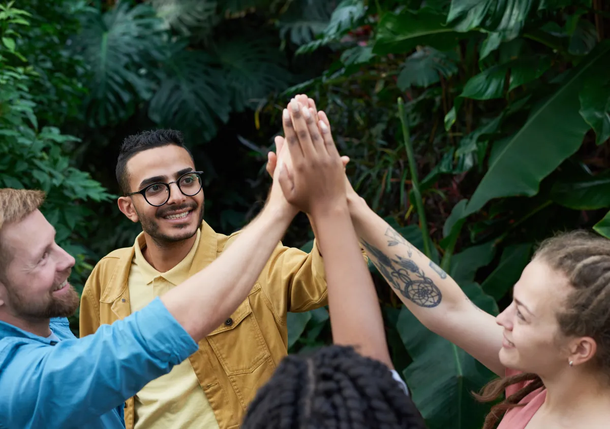 Four young adults with right hands raised forming a group high-five; all different ethnicities; one woman having tattoos on her arm, one man with glasses.