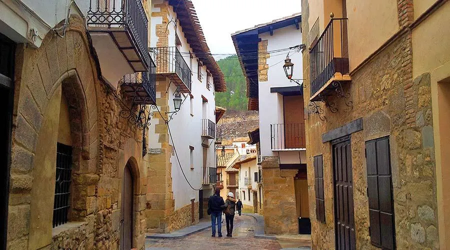 A street view of Rubielos de Mora, a medieval village nestled in the Sierra de Gúdar, Teruel, Spain. The image showcases the town's well-preserved stone architecture, including the iconic Portal de San Antonio and the Ex Colegiata de Santa María la Mayor, set against a backdrop of rolling hills and clear blue skies. Narrow cobblestone streets wind through historic buildings adorned with wooden balconies and wrought-iron details, embodying the charm that earned Rubielos de Mora the Europa Nostra award for heritage conservation. This picturesque setting reflects the town's commitment to the Cittaslow movement, promoting a tranquil and culturally rich lifestyle.