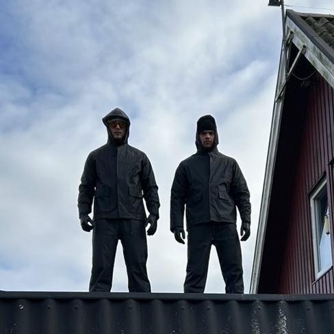 Medarbetare från Spindelmännen i arbetskläder framför en ljus fasad, fotografi i naturligt dagsljus, fokus på professionell utrustning och vänligt kroppsspråk för att kommunicera trygghet och kunnande.