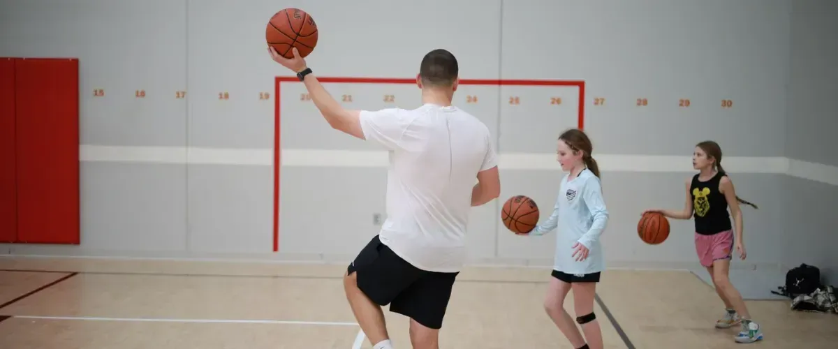 Coach leading basketball drill with two young girls practicing dribbling.