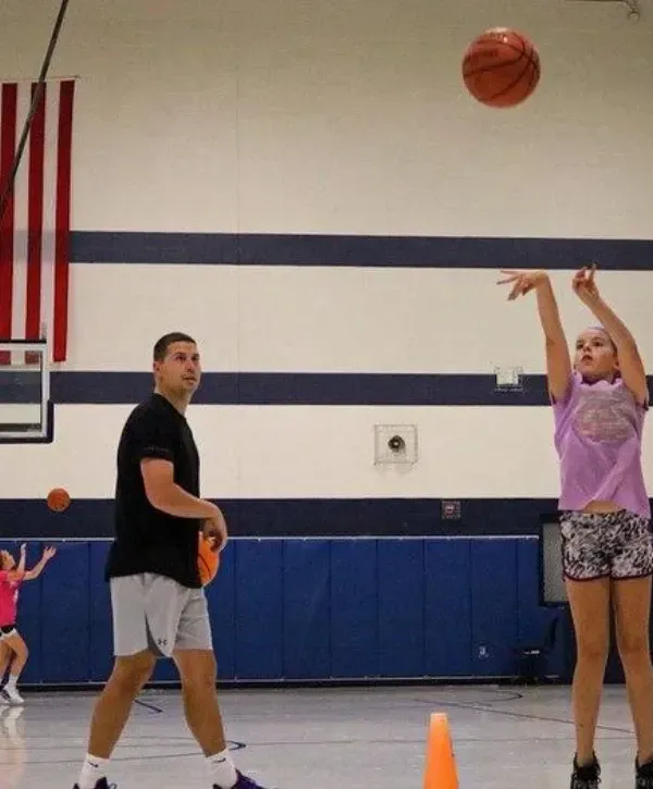 Young girl practicing basketball shooting form with a coach supervising in a gym.