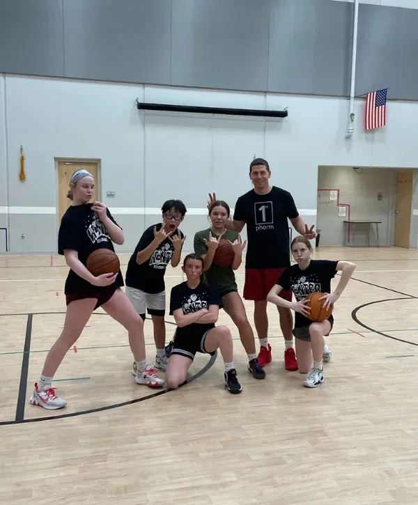 Youth basketball team posing with their coach on an indoor court, holding basketballs and making playful gestures.