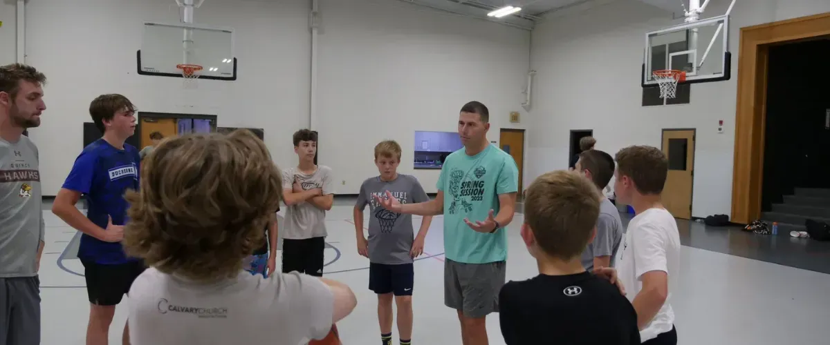 Basketball coach speaking to a group of youth players gathered in a huddle on an indoor court.