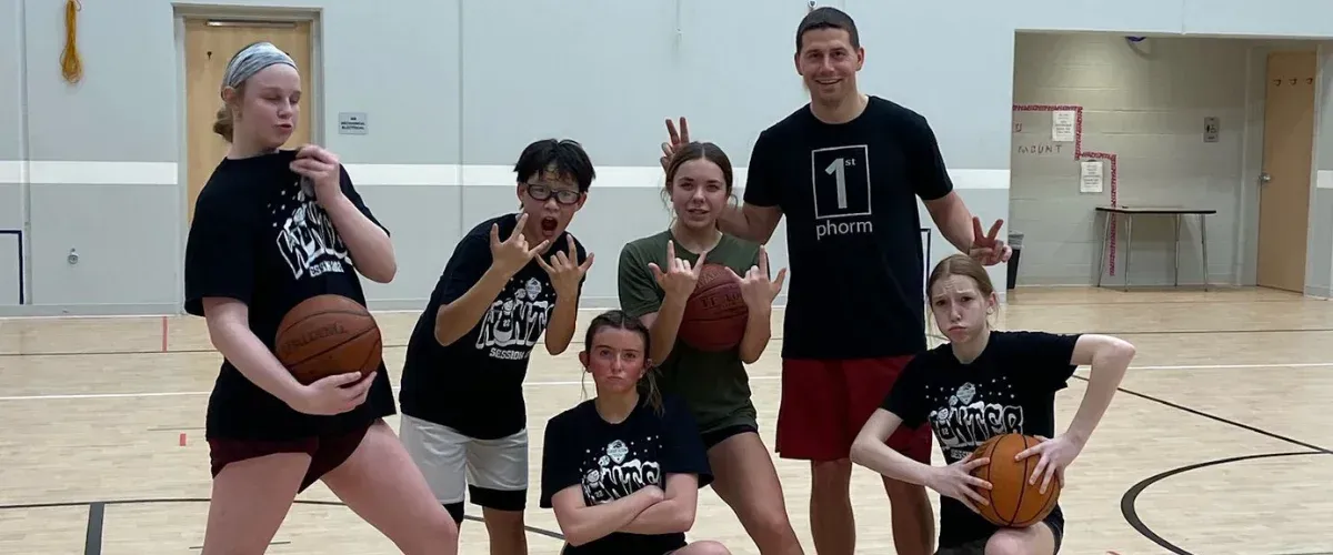 A male basketball coach in a black shirt holds a basketball while instructing young players on the court.