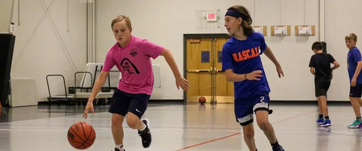 Two boys actively dribble basketballs on a shiny gymnasium court during a game or practice, with others in the background.
