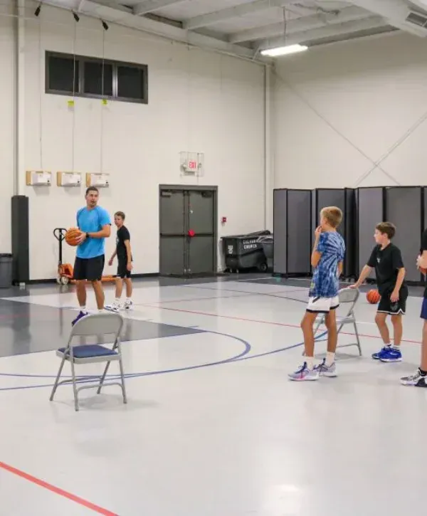 A basketball coach holding a ball instructs three young boys on a court during a clinic near chairs.