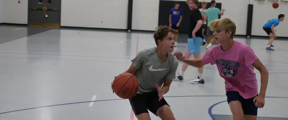 A teen boy in a gray Nike shirt dribbles a basketball on a gym court while another boy in pink defends.