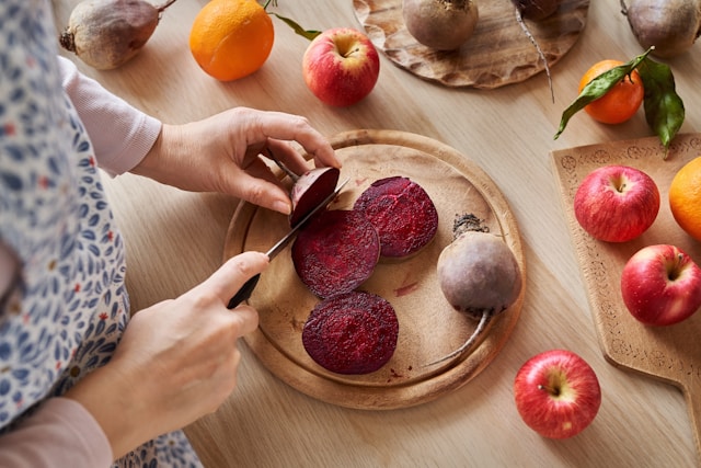Hands slicing fresh beets on a wooden cutting board surrounded by apples and oranges, representing heart-healthy whole foods relevant to managing MS alongside other health conditions.