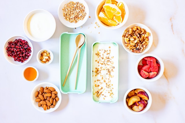 Yogurt, oats, walnuts, pomegranate seeds, citrus, almonds, and honey arranged in small white bowls on a marble surface with wooden spoons, representing anti-inflammatory whole foods for MS nutrition.