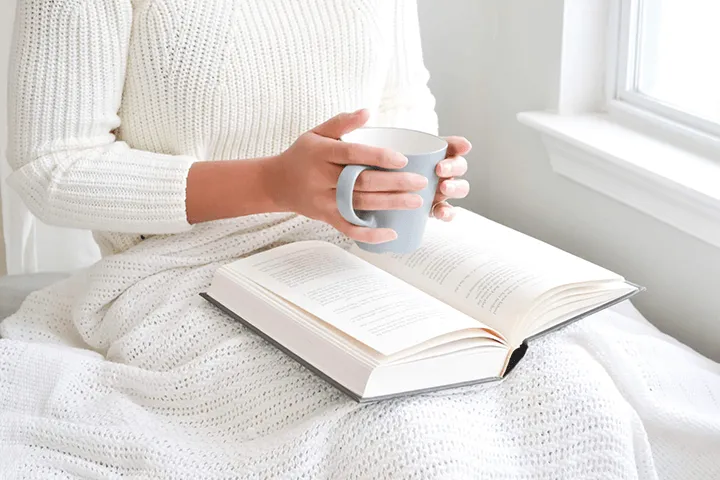 A person in a white sweater holding a warm mug beside an open book in soft window light, representing calm and reassurance for someone newly diagnosed with MS.