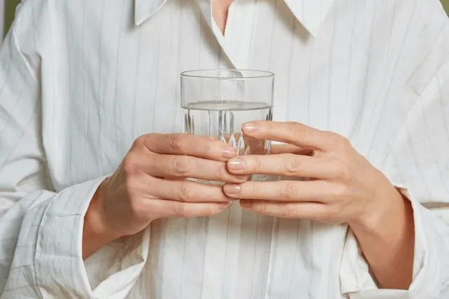 A person in a white collared shirt holds a clear glass of water with both hands.