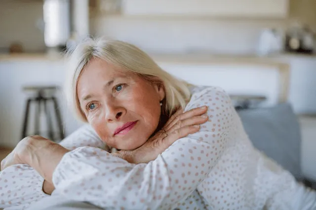 A woman with blonde hair rests her head on her arms, looking off to the side with a thoughtful expression.
