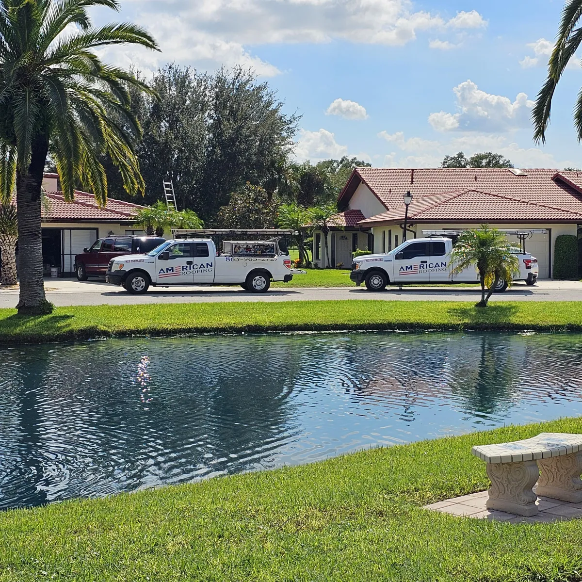 American Roofing FL trucks by a neighborhood pond with palm trees and tile-roof homes.