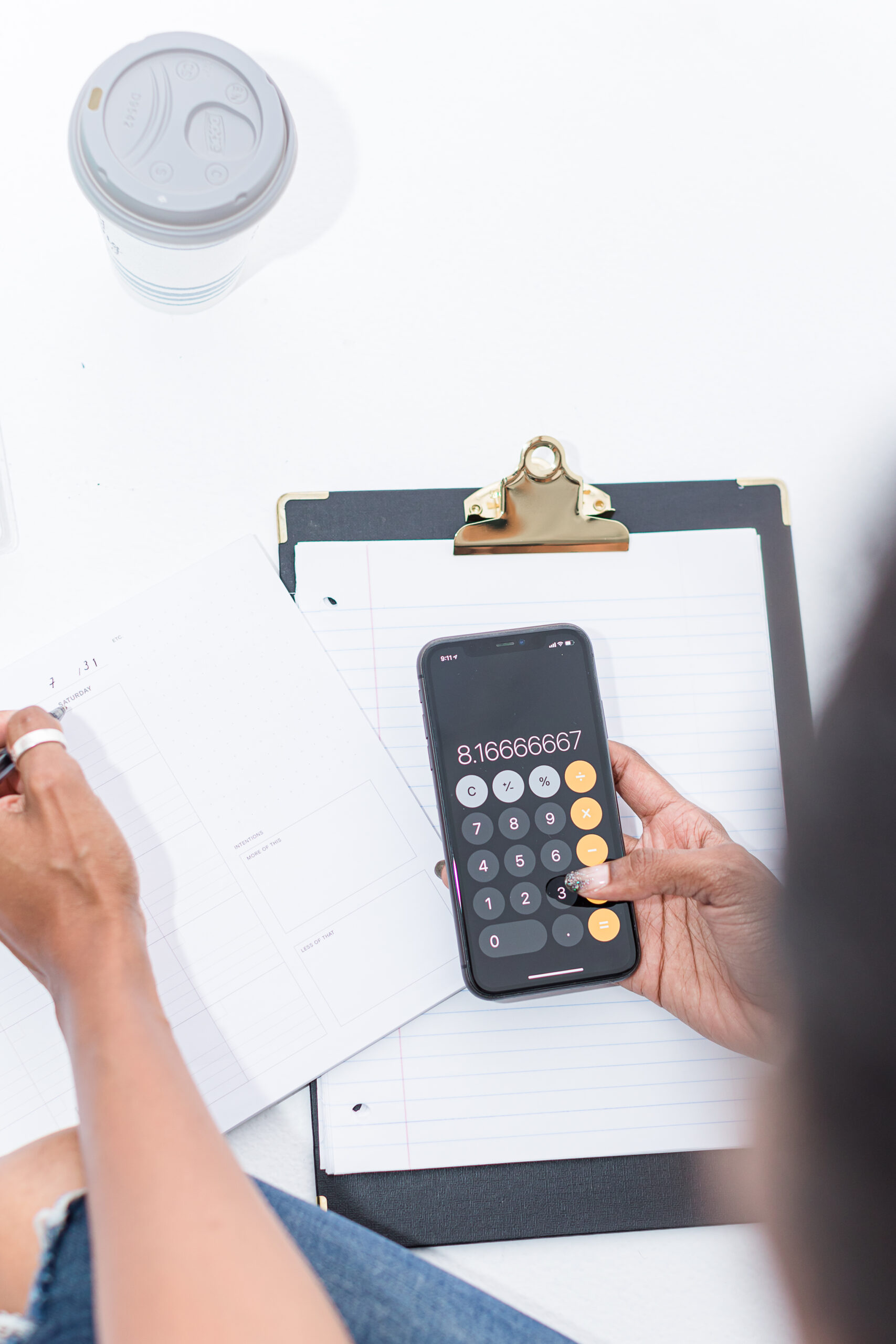 Overhead view of hands planning finances: writing in an open planner while holding a phone with a calculator app; clipboard and coffee cup nearby.