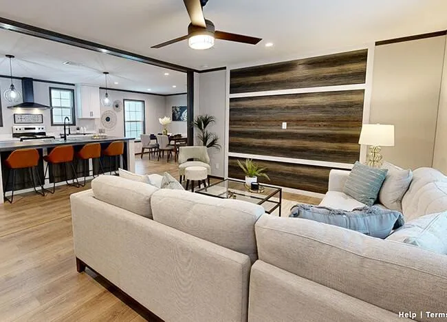 A modern open-concept living space in a Houston Embra Manufacture Home features a white sectional sofa facing a wall-mounted TV, adjacent to a kitchen with a breakfast bar and stools, and a dining area. The room is complete with a ceiling fan and elegant wooden flooring.