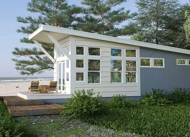 A modern, small house with large windows, a sloped roof, and a wooden deck with chairs, surrounded by greenery. The house is situated near a sandy beach with trees in the background.