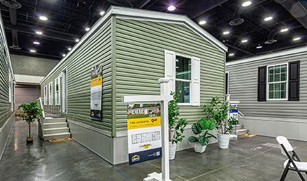 A green manufactured home display inside an exhibition hall, surrounded by plants. Informational posters and banners are visible, along with a sign featuring floor plans and details about The Avenue.