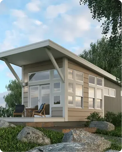 A modern tiny house with large windows and a slanted roof sits on a wooden deck. Two chairs and a small table are placed outside. Trees and large rocks surround this charming park home.