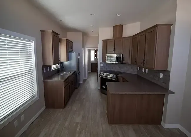 A narrow, modern kitchen with wooden cabinets, stainless steel appliances, and a window with blinds, featuring a tiled backsplash and light hardwood floors.