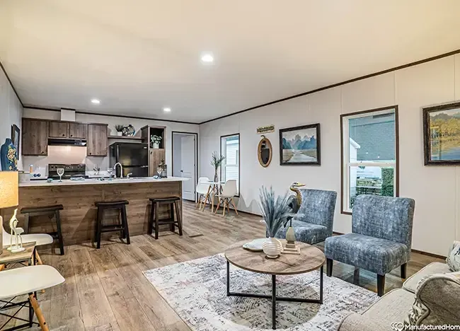 A modern living area in a Houston PHX 66 Manufactured Home boasts wooden flooring, a kitchen island with three stools, and a seating area with two blue chairs and a round coffee table. The walls feature paintings while windows provide natural light, creating an inviting atmosphere.