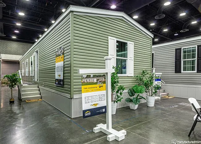 A green manufactured home with white trim on display at an indoor exhibition. The home features steps leading to the door, plants, and informational signs on display.