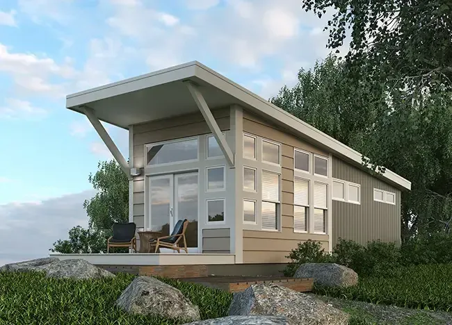 Modern tiny house by J Houston Park Homes with a sloping roof, large windows, and a small deck furnished with two chairs and a table, all surrounded by lush greenery and rocks.