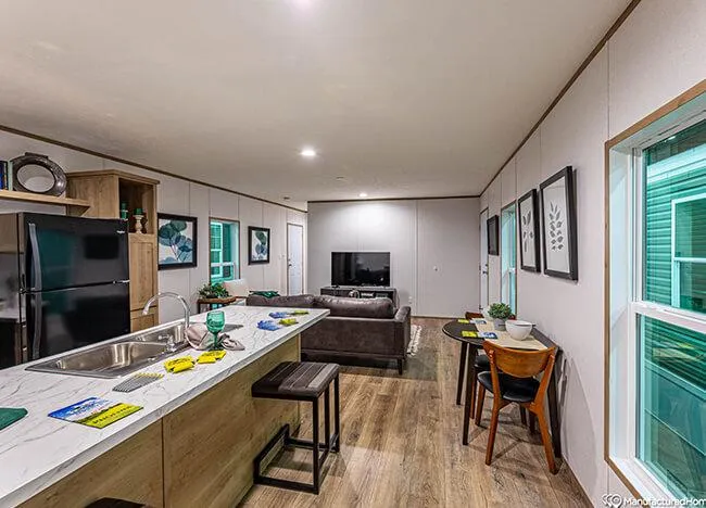 A modern, open-concept living space featuring a kitchen with an island, a dining area, and a living room with a sofa and wall-mounted TV. The decor includes framed art and various household items.