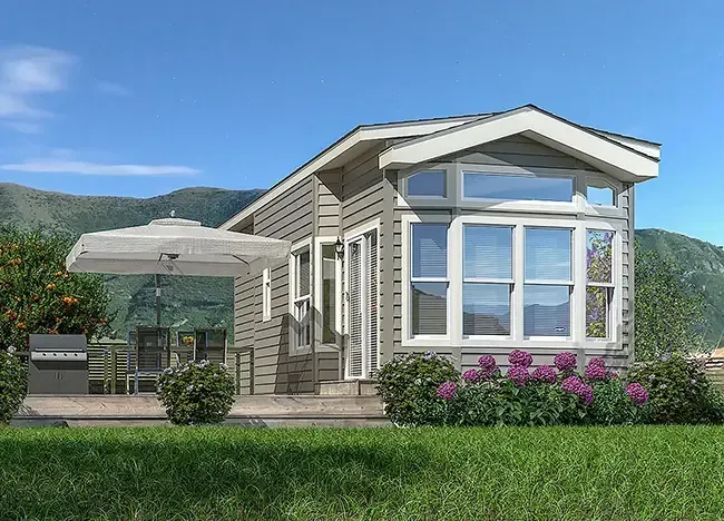 A small beige J Houston Manufactured Homes house with large windows and a patio featuring an outdoor umbrella, grill, and dining table set, set against a backdrop of green hills and a blue sky.