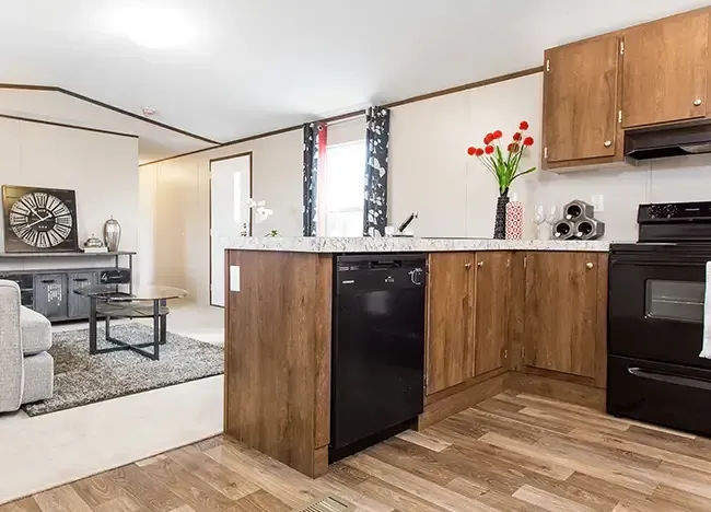 A modern kitchen with wooden cabinets, black appliances, and granite countertops. A vase with red tulips is on the counter. The living area has a sofa, round coffee table, and a rug.