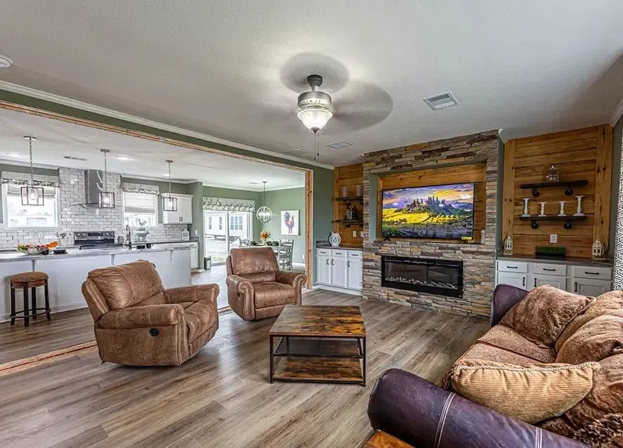 Spacious living room with two brown recliners, a brown couch, a wooden coffee table, a stone fireplace with a TV above it, and an open view of the kitchen and dining area by JHouston Properties.