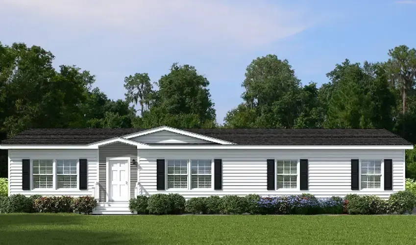 A single-story manufactured home with brown siding, white trim, and a front porch, set in a grassy area surrounded by trees under a clear blue sky; this Houston Apex property offers serene living at its finest.