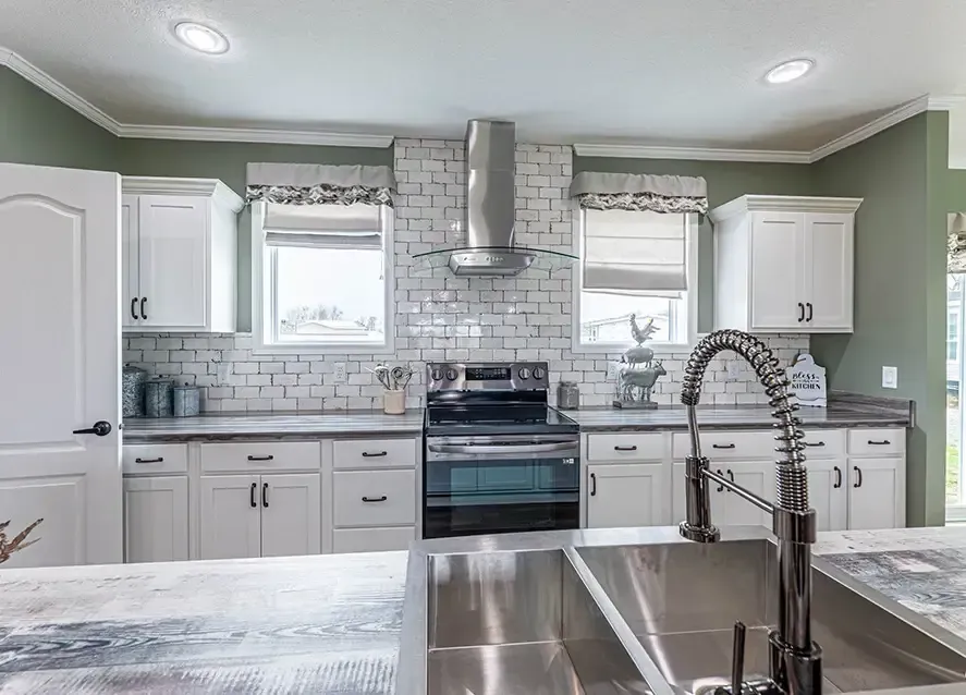 A modern kitchen with green walls features white cabinets, a stainless steel oven and range hood, a dual sink on a gray countertop, and a white tile backsplash. Two small windows are above the stove.