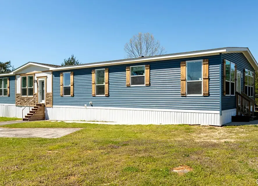 A white, single-story manufactured home with black shutters and a sloping roof stands in front of a lush, green background with trees. Shrubs and plants line the front of the Houston Brantley house.