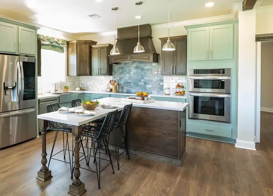 Modern kitchen with light green cabinets, stainless steel appliances, and a large island with seating. The backsplash is a mix of blue tiles, and the flooring is wood. Pendant lights hang above the island.