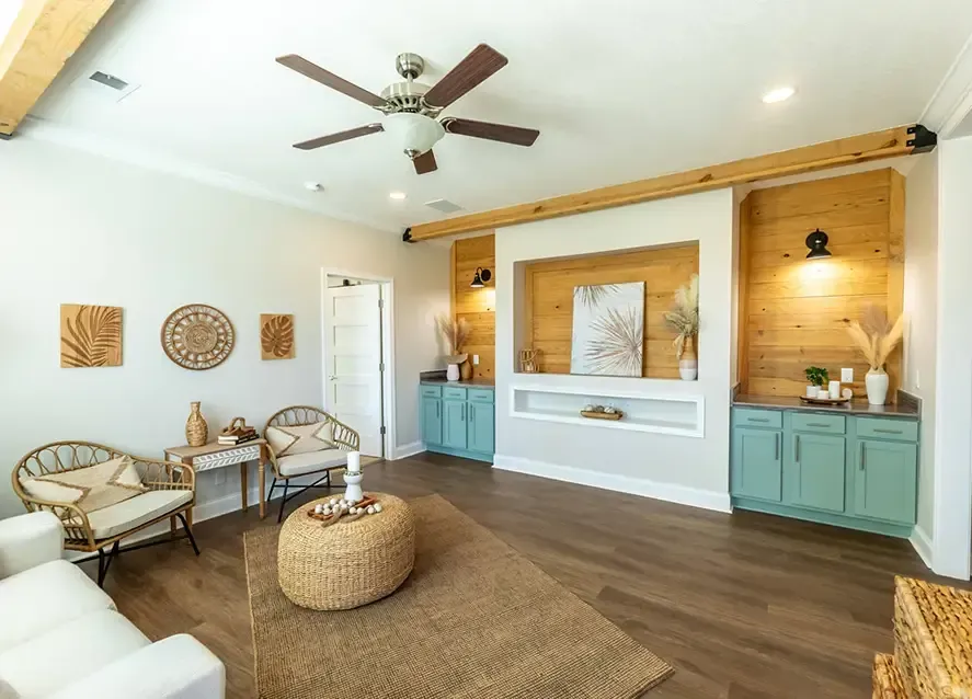 A modern living room in a manufactured home boasts wooden floors, a ceiling fan, light blue cabinets, wicker furniture, and decorative wall art. Two armchairs and a table are set against one wall, with a round ottoman in the center.