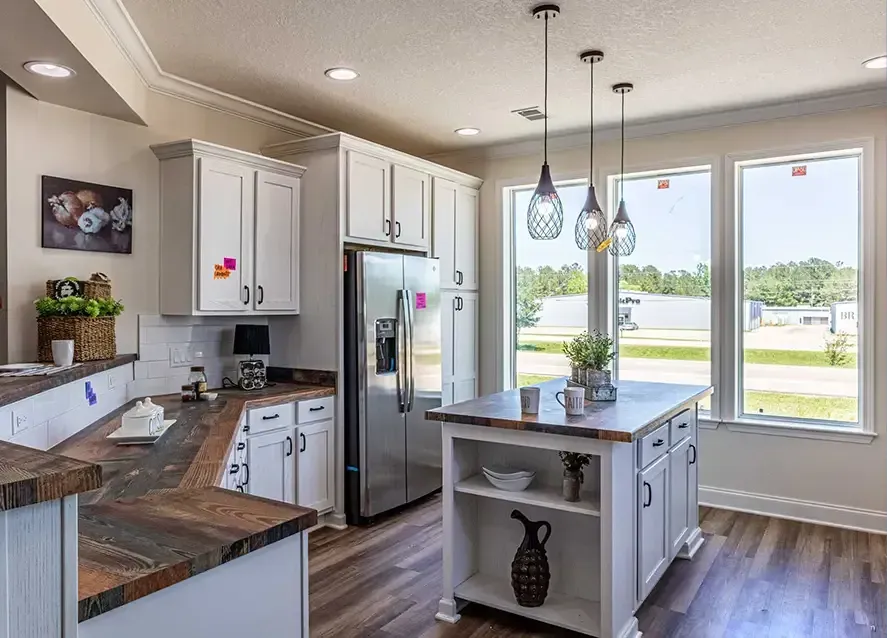 A modern kitchen with white cabinets, stainless steel appliances, a wooden countertop island, pendant lighting, and large windows providing natural light. There are decorative items on the shelves and counter.