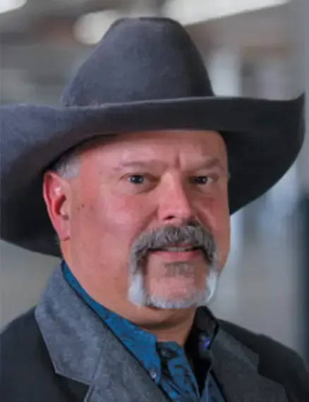 A lender for Houston financing, a man with a gray mustache and goatee, is wearing a large gray cowboy hat and a dark jacket over a blue shirt. He is looking into the camera with a neutral expression.