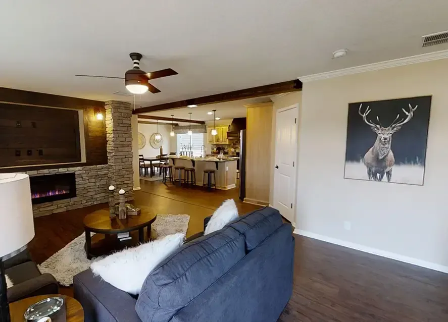 Living room with a grey couch, stone fireplace with TV above, and view into kitchen. Ceiling fan, wooden floors, and deer painting on the wall. Open layout with modern decor in this Houston Apex Manufactured Home.