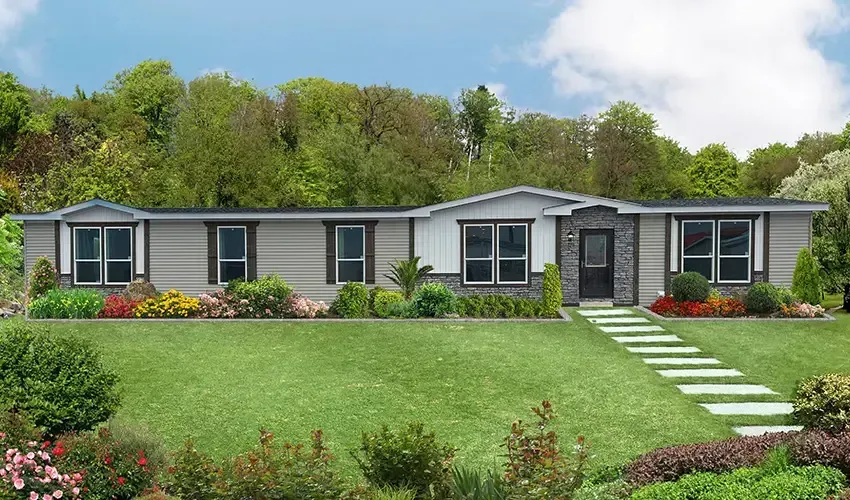 A single-story manufactured home with a front porch and steps set in a grassy area with trees and flower beds nearby under a partly cloudy sky.