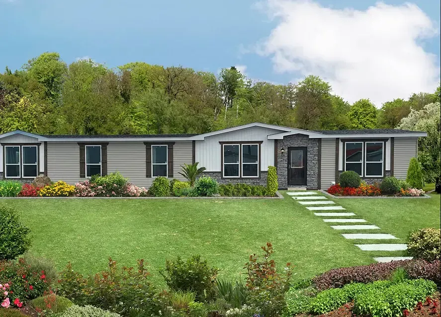 A modern, single-story Houston Apex Manufactured Home features a grey and white exterior, large windows, a well-manicured lawn, a stone pathway, and a landscaped garden set against a backdrop of trees and a partly cloudy sky.