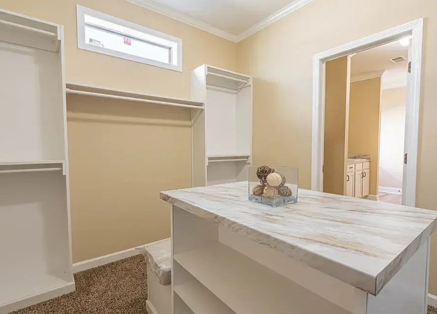 A walk-in closet with beige walls, white shelves, and a center island featuring a marbled countertop adorned with decorative items. An adjoining bathroom with a sink is visible through the open door of this Houston Woodland manufactured home.