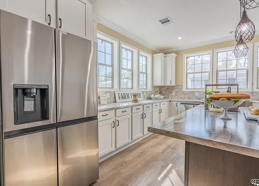 A modern kitchen with stainless steel appliances, white cabinets, and wooden flooring. A kitchen island features a decorative cake stand with fruit. Sunlight streams through multiple windows.