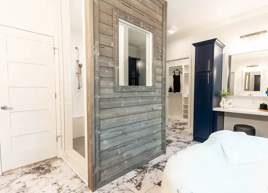 Modern bathroom with marble floor, centered wooden partition featuring a mirror, shower area to the left, dark blue vanity on the right, and a dressing area in the background.