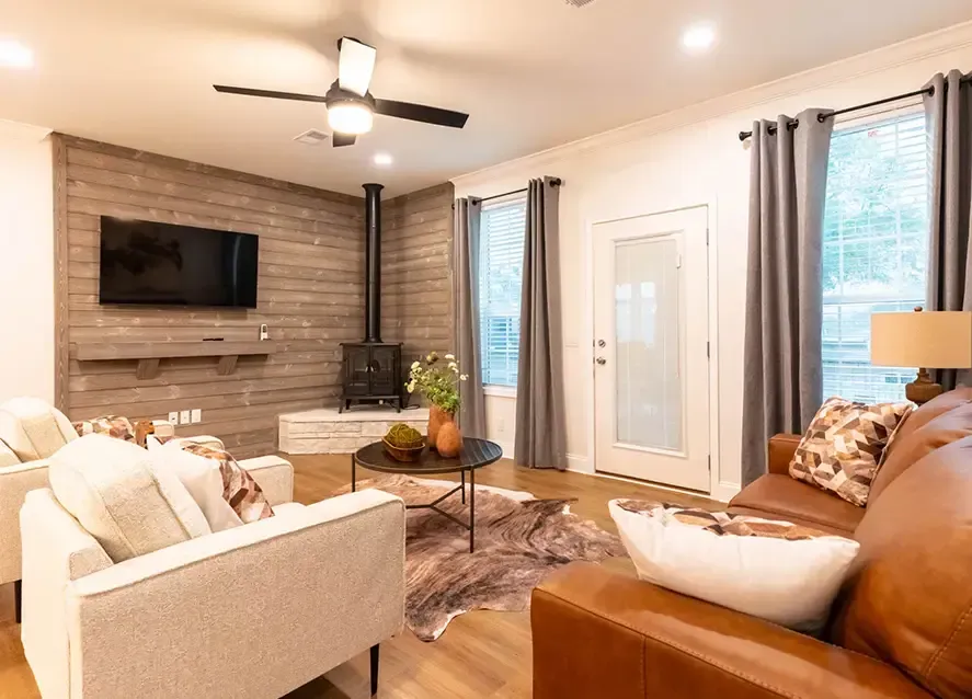 A cozy living room in a Houston Shoreline Manufactured Home features a brown leather couch, two white armchairs, a round coffee table, and a wall-mounted TV. A wood-burning stove adds warmth, while a ceiling fan and large windows with gray curtains enhance the inviting space.