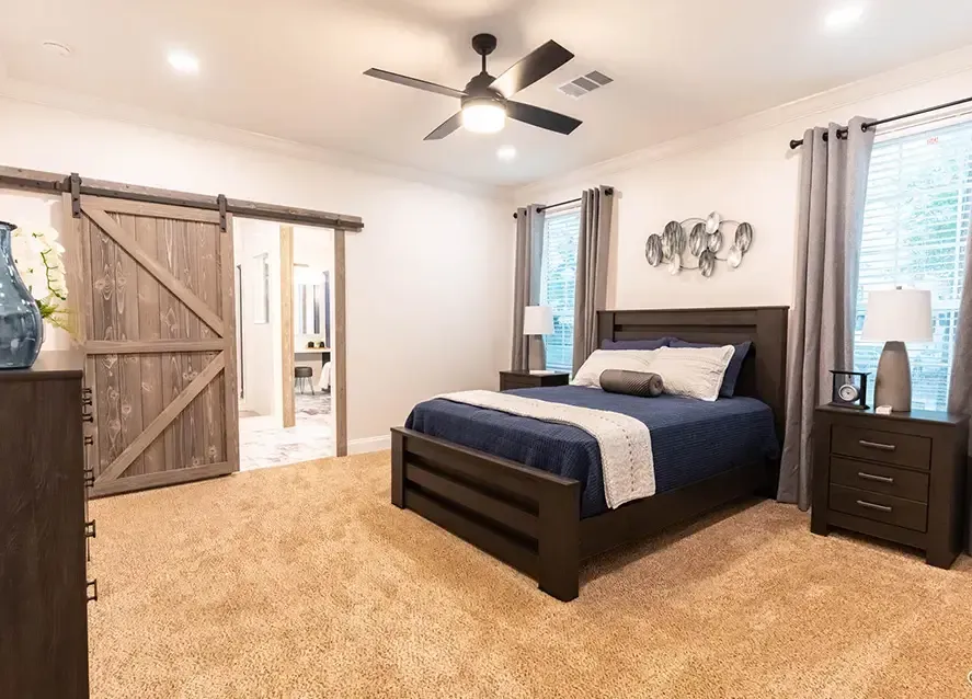 A modern bedroom in a Houston Shoreline Manufactured Home features a large bed with navy bedding, two nightstands with lamps, a ceiling fan, carpeted floor, barn-style sliding door, and two windows with gray curtains.